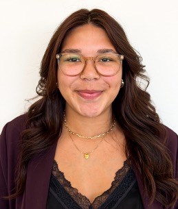 Photo of smiling female. She has long dark hair, is wearing glasses and a black professional top with maroon blazer.